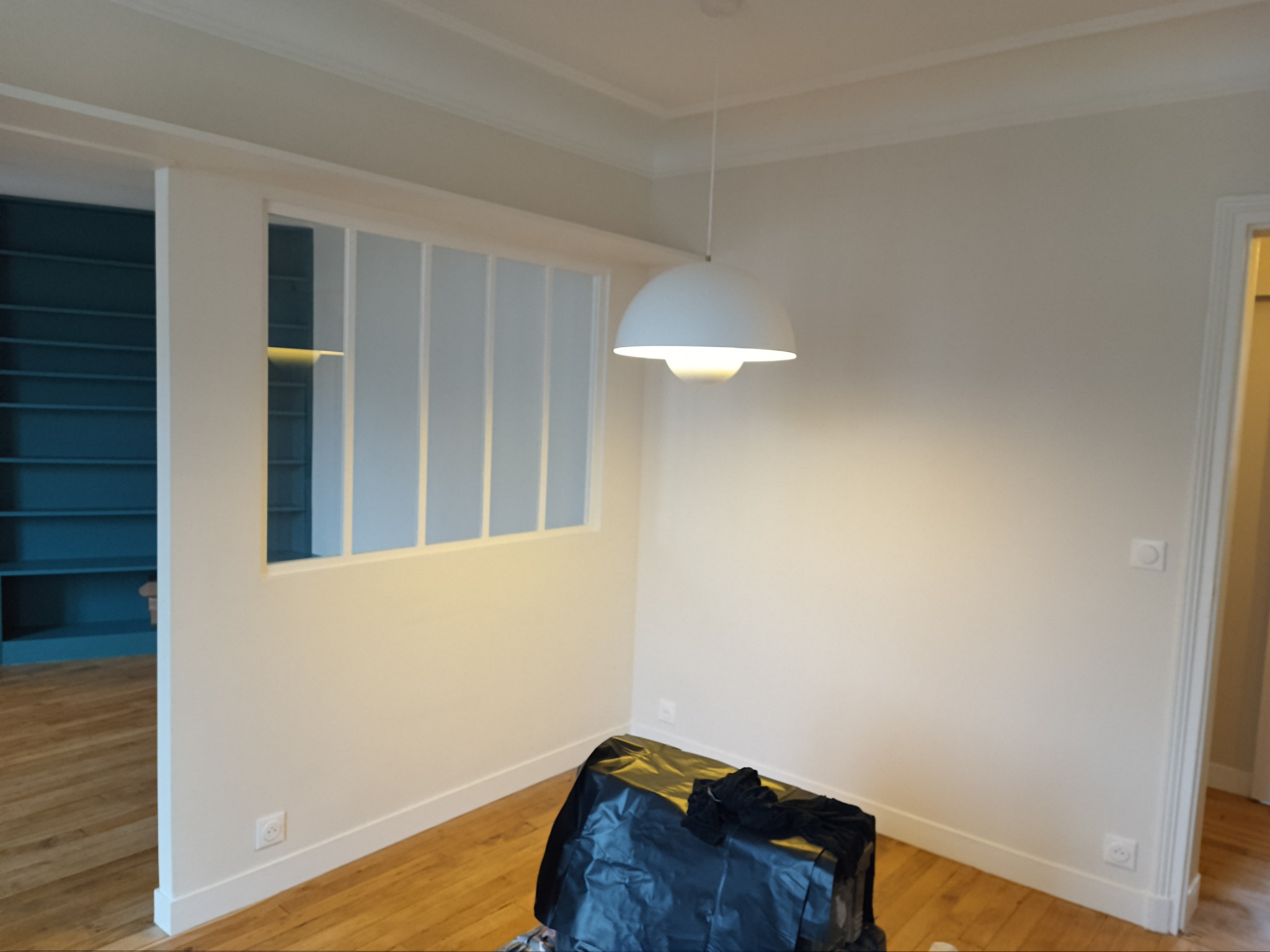 Pokój w trakcie przygotowań do prac wykończeniowych: ściany pomalowane na biało, drewniana podłoga zabezpieczona folią, wisząca lampa w stylu skandynawskim, widoczny fragment niebieskiego regału...