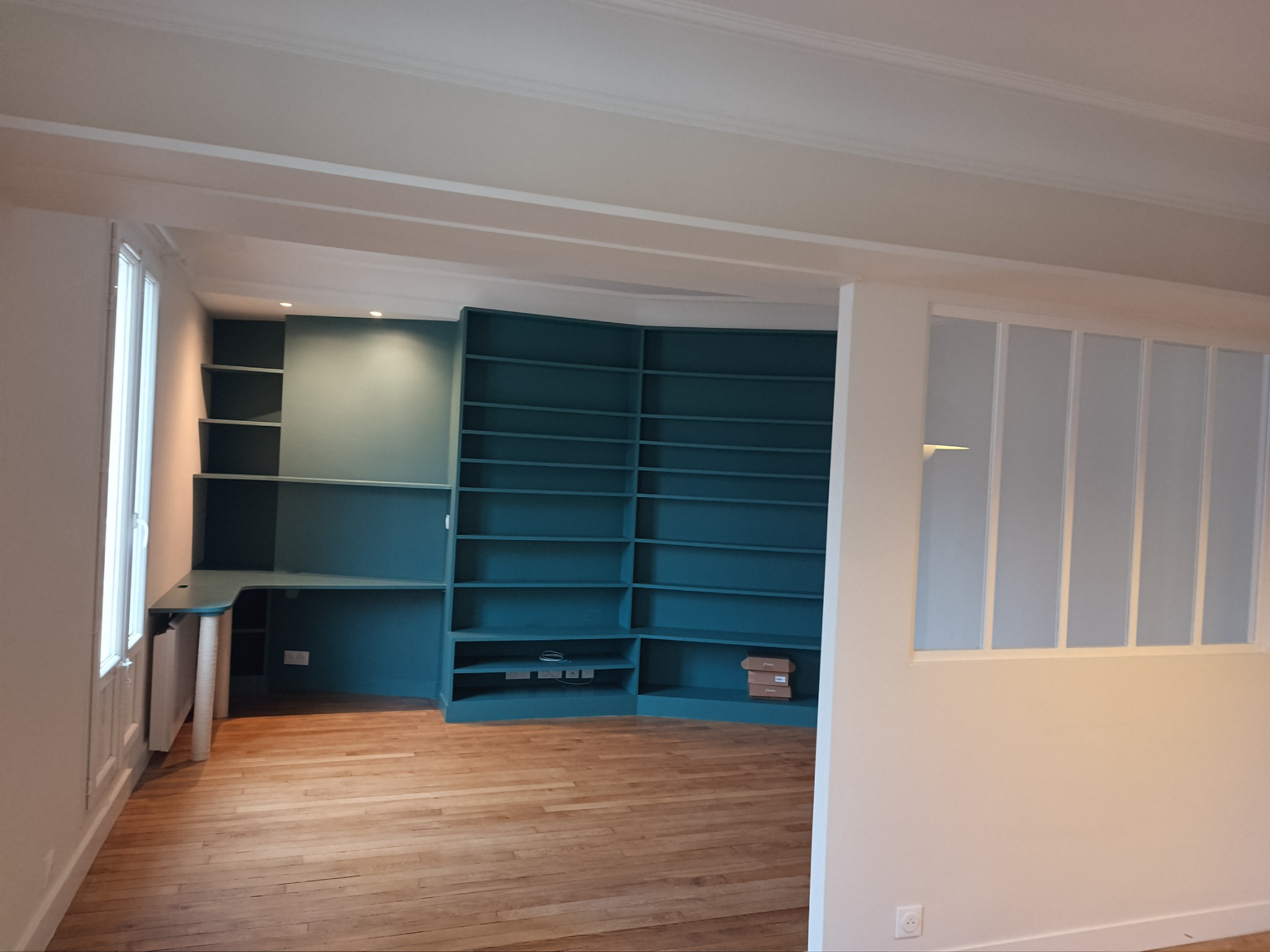 Turkusowa zabudowa meblowa z regałami i biurkiem w jasnym pokoju z parkietem, widok na ścianę z oknem i grzejnikiem.