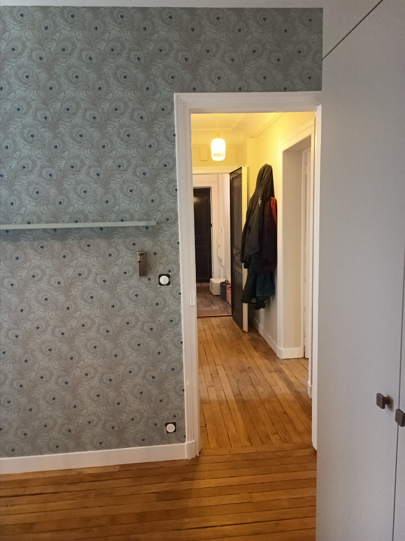 Korytarz po remoncie z wzorzystą tapetą w odcieniach szarości i błękitu z motywem pawich piór, drewnianą podłogą i białymi drzwiami na końcu korytarza.