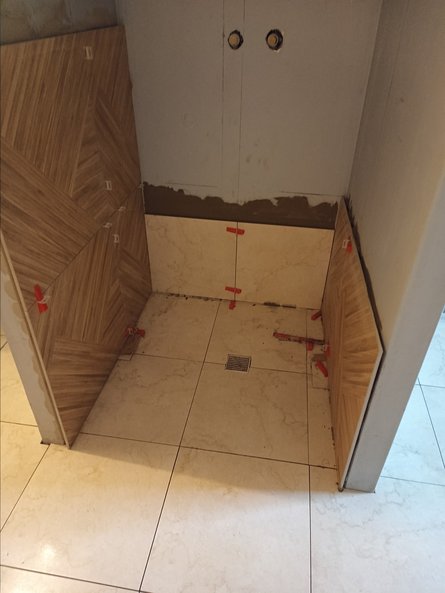 Fragment łazienki w trakcie układania płytek pod prysznicem, widoczne płytki imitujące drewno na ścianie i marmur na podłodze, z czerwonymi krzyżykami dystansowymi i odpływem liniowym.