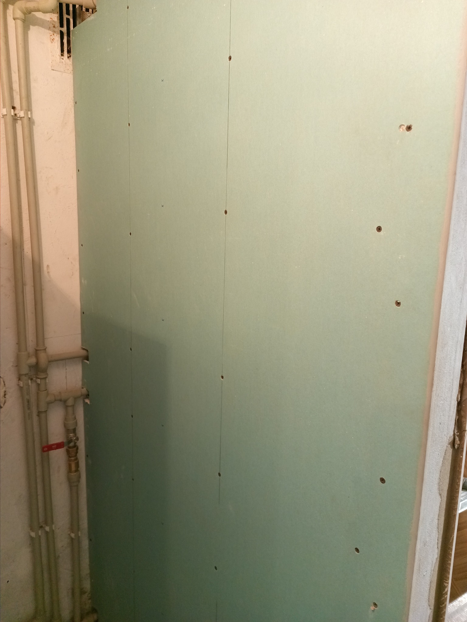 Ściana z zielonej płyty kartonowo-gipsowej, przymocowana do ramy, widoczne śruby mocujące, obok rury instalacyjne.