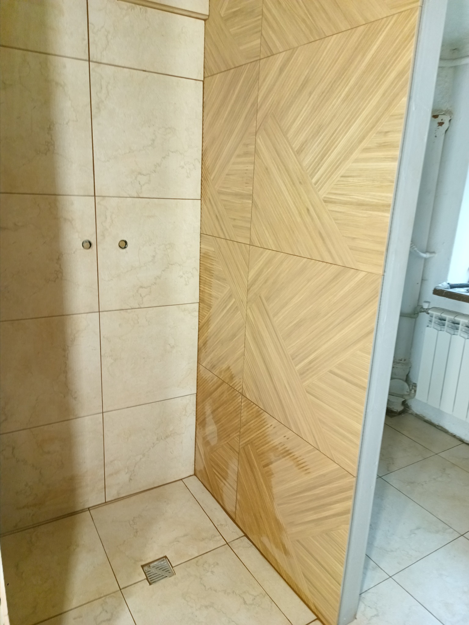 Wykończona kabina prysznicowa z beżowymi płytkami imitującymi marmur oraz drewnopodobnymi płytkami ułożonymi w geometryczny wzór na ścianie bocznej, widoczny odpływ liniowy w podłodze.