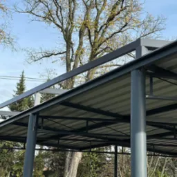 Zadaszenie metalowe z szarej blachy trapezowej, wsparte na okrągłych, szarych słupach. Widoczne drzewa i błękitne niebo w tle.