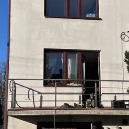 Balkon z nową, metalową balustradą z poziomych prętów. Widoczne narzędzia i skrzynka narzędziowa wskazują na trwający montaż. Jasne tło budynku i niebieskie niebo.