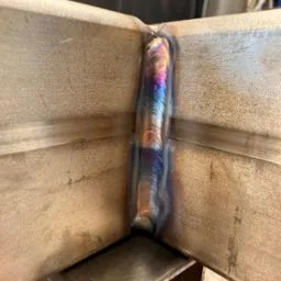 Spoina łącząca dwa elementy metalowe pod kątem prostym, z widocznymi tęczowymi przebarwieniami po spawaniu. Detal wykonania, zbliżenie na łączenie.