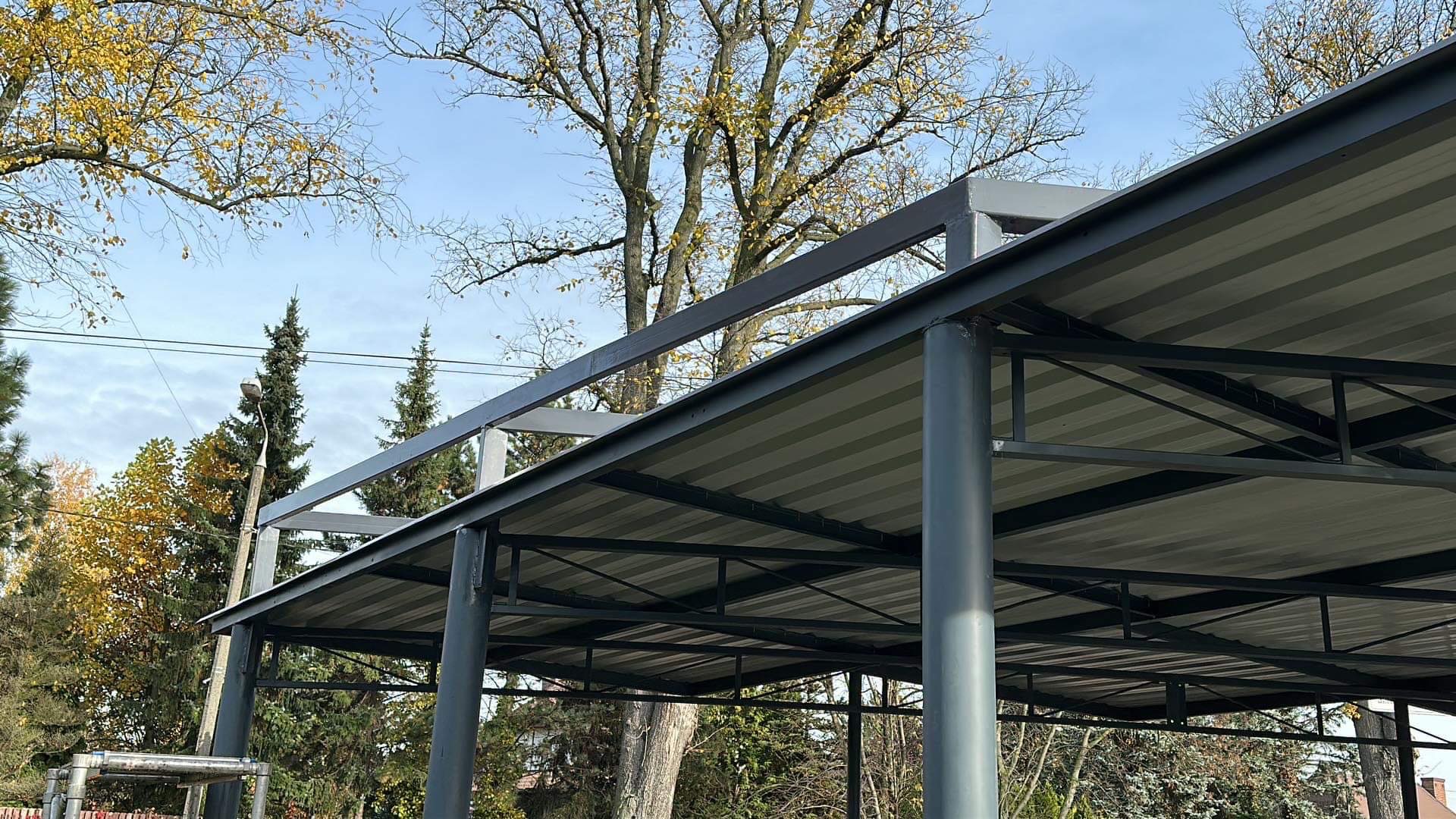 Zadaszenie metalowe z szarej blachy trapezowej, wsparte na okrągłych, szarych słupach. Widoczne drzewa i błękitne niebo w tle.
