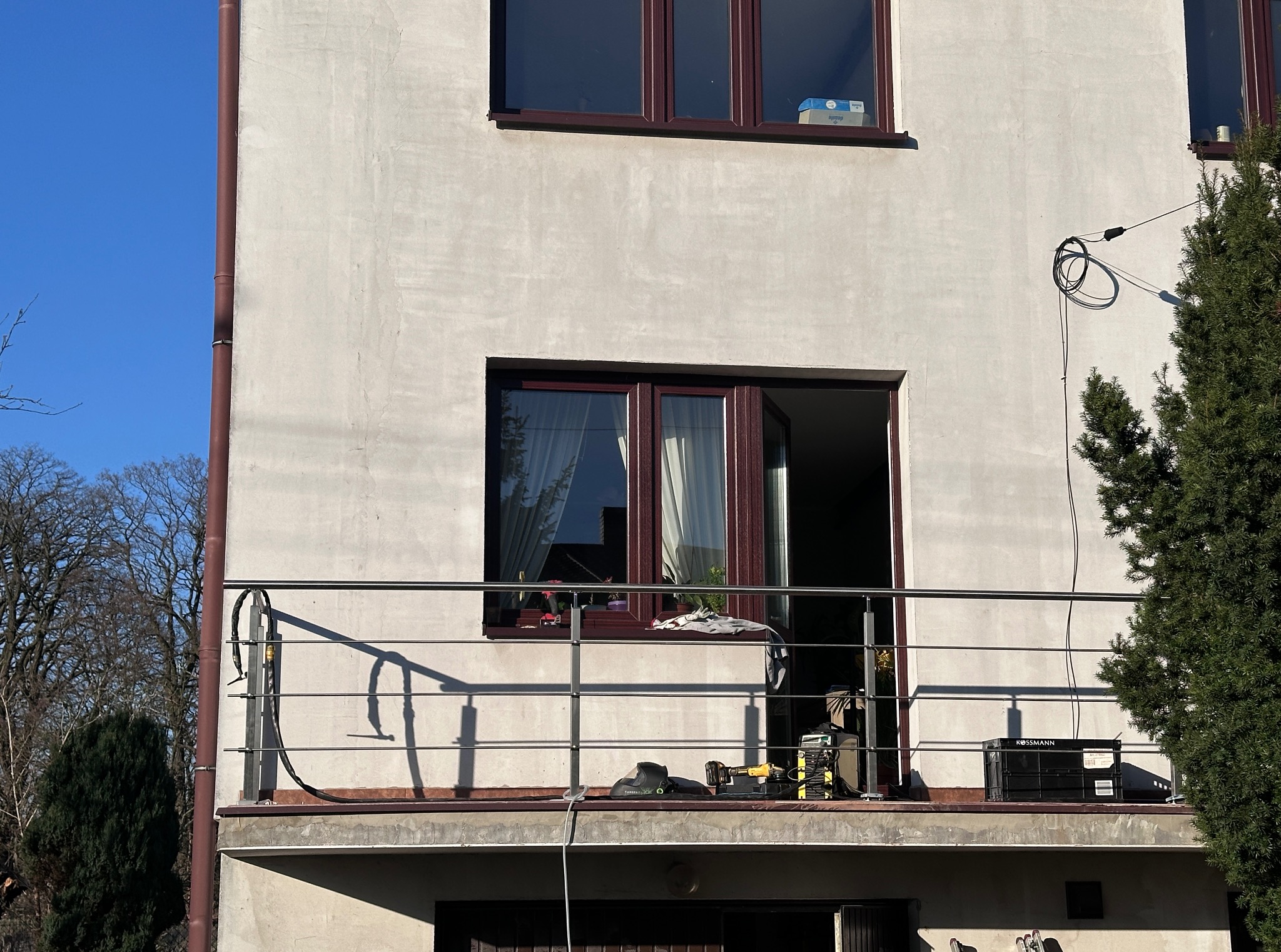 Balkon z nową, metalową balustradą z poziomych prętów. Widoczne narzędzia i skrzynka narzędziowa wskazują na trwający montaż. Jasne tło budynku i niebieskie niebo.