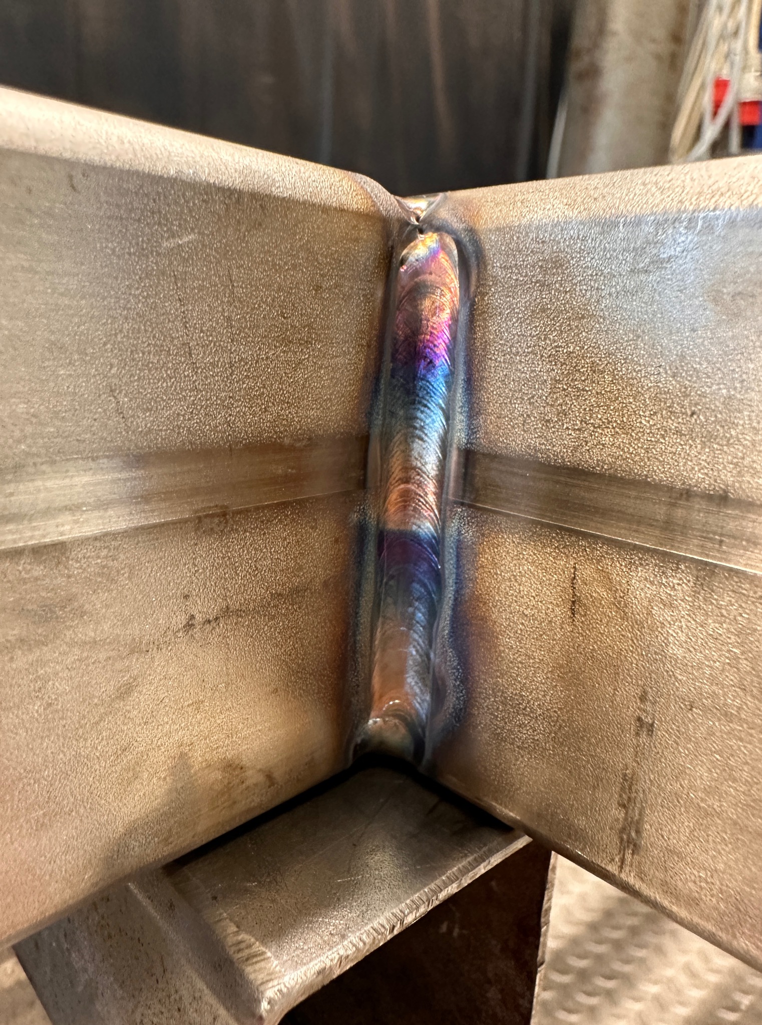 Spoina łącząca dwa elementy metalowe pod kątem prostym, z widocznymi tęczowymi przebarwieniami po spawaniu. Detal wykonania, zbliżenie na łączenie.