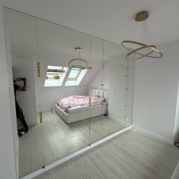 Jacek szklo - Duża, lustrzana szafa wnękowa z złotymi mocowaniami, odbijająca wnętrze sypialni z łóżkiem i oknem dachowym; widoczne odbicie fotografa.