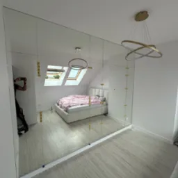 Duża, lustrzana szafa wnękowa z złotymi mocowaniami, odbijająca wnętrze sypialni z łóżkiem i oknem dachowym; widoczne odbicie fotografa.