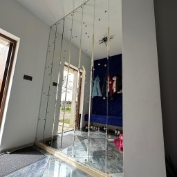 Jacek szklo - Montaż lustrzanej ściany z pionowych paneli, zabezpieczonych taśmą, w jasnym przedpokoju z widocznym wejściem i elementami garderoby w odbiciu.