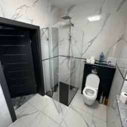 Nowoczesna łazienka z kabiną prysznicową ze szklanymi ścianami, czarną ramą i drzwiami otwieranymi na zewnątrz, białe marmurowe płytki na ścianach i podłodze, czarne drzwi wejściowe.