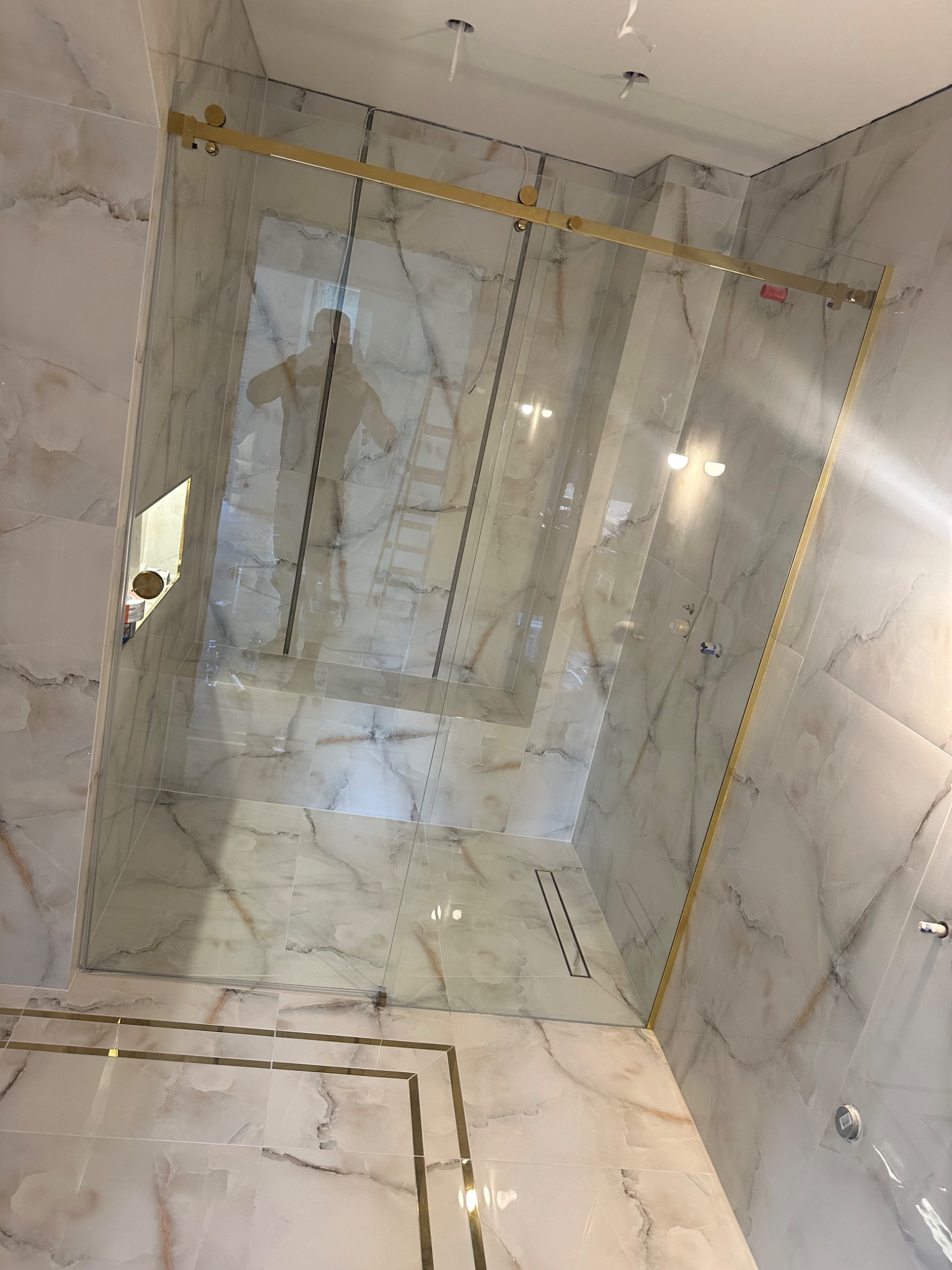 Szklana kabina prysznicowa z przesuwnymi drzwiami i złotymi okuciami, widoczna w łazience wyłożonej marmurowymi płytkami, odbicie osoby na drabinie w szkle.