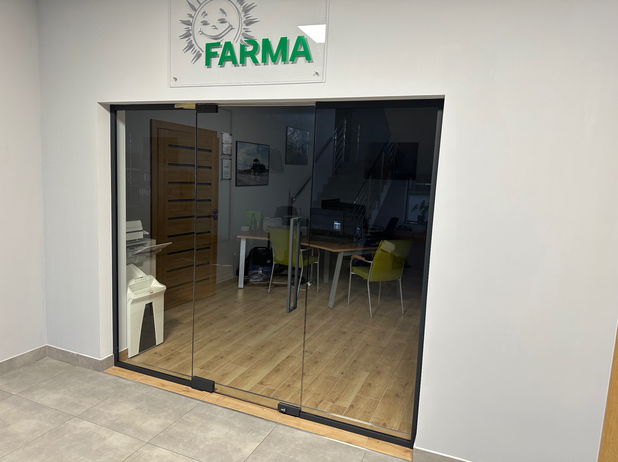 Przesuwne drzwi szklane z czarną ramą w biurze, widok na wnętrze z biurkami i krzesłami, logo firmy 'FARMA' nad wejściem.