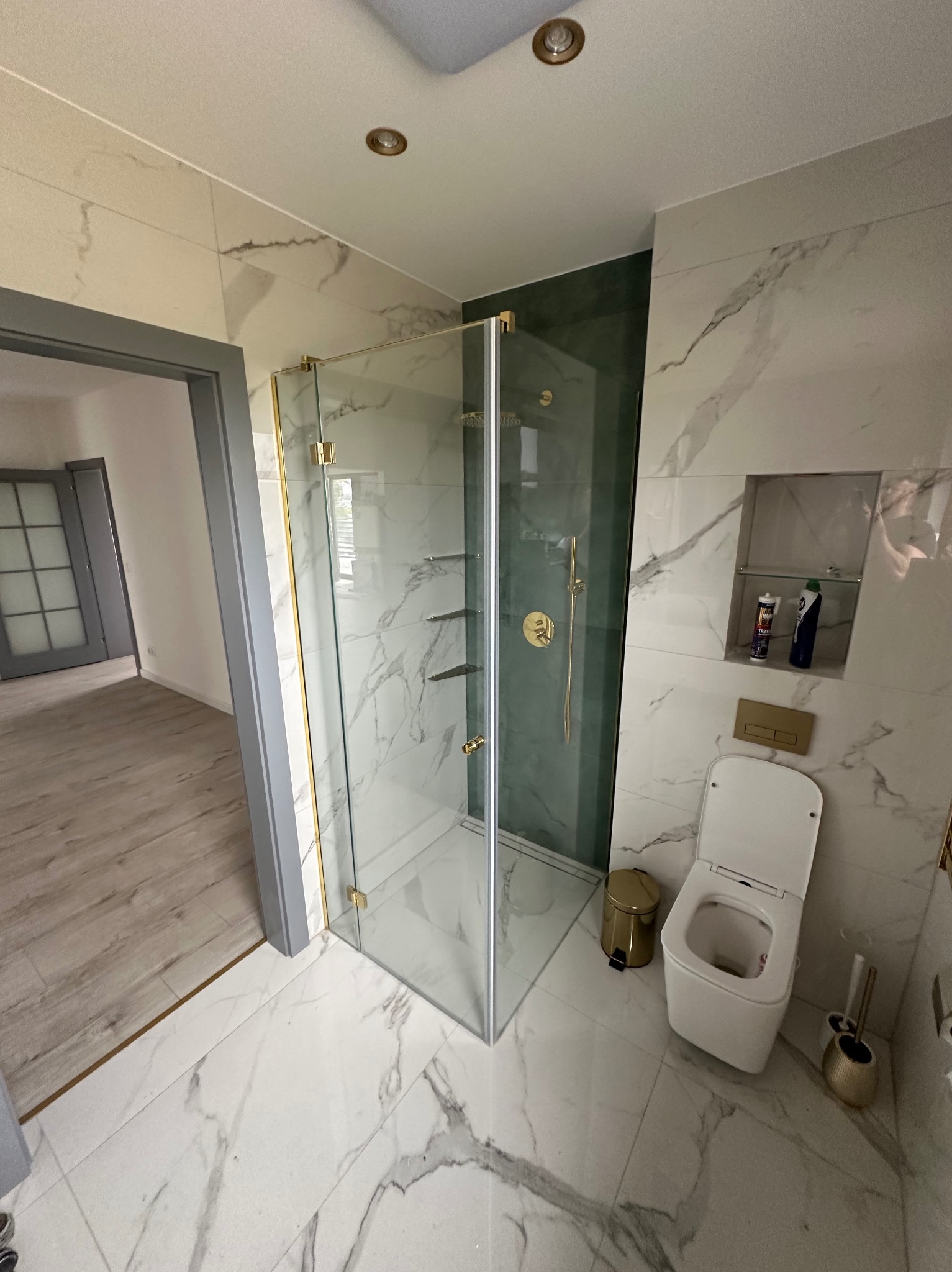 Nowoczesna łazienka z kabiną prysznicową ze szkła hartowanego z złotymi okuciami, marmurowe płytki na ścianach i podłodze, widoczna toaleta i akcesoria łazienkowe w kolorze złotym.