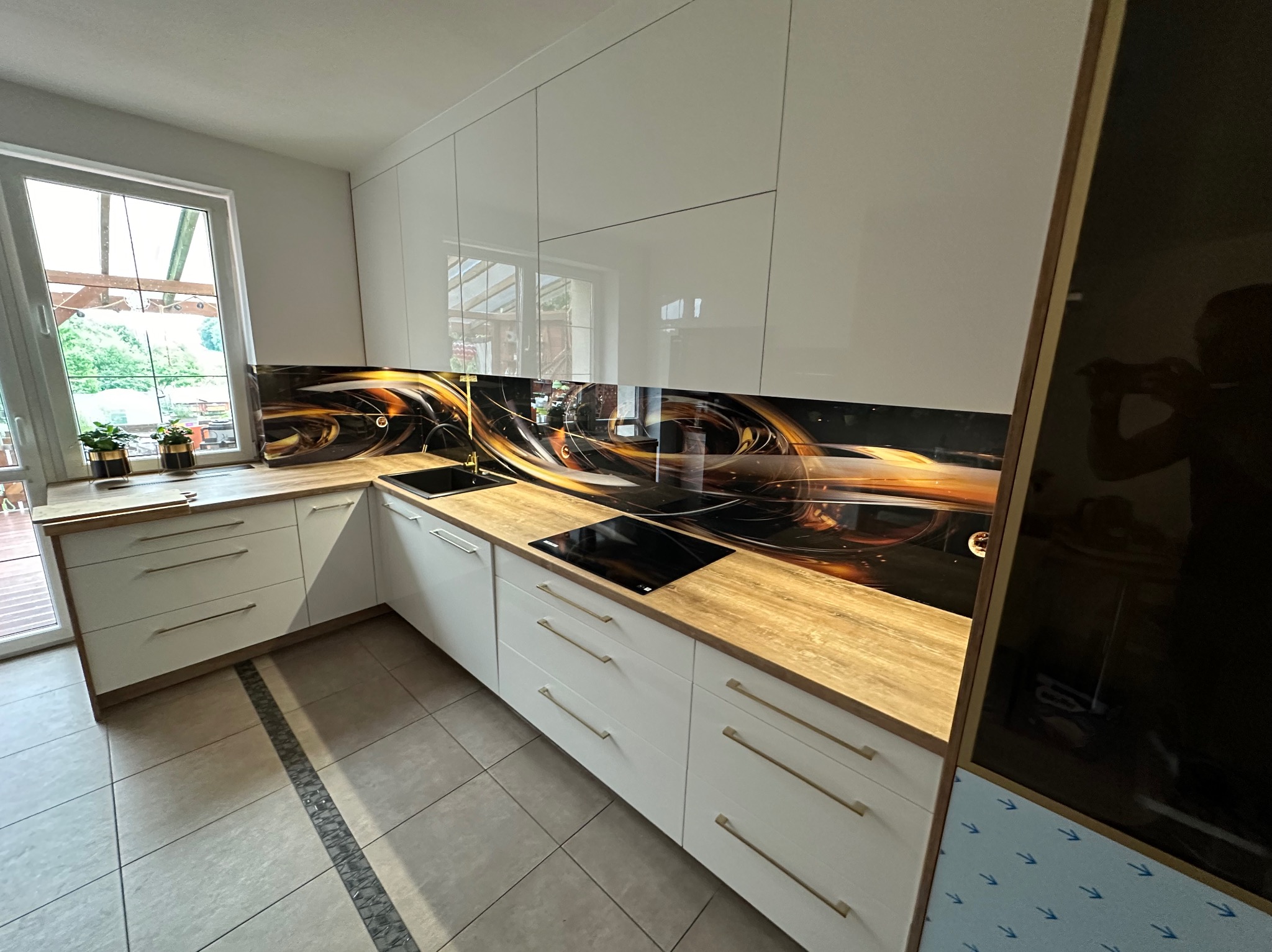 Nowoczesna kuchnia z białymi szafkami i blatem imitującym drewno, ozdobiona panelem szklanym z abstrakcyjnym, złotym wzorem na ścianie nad blatem.