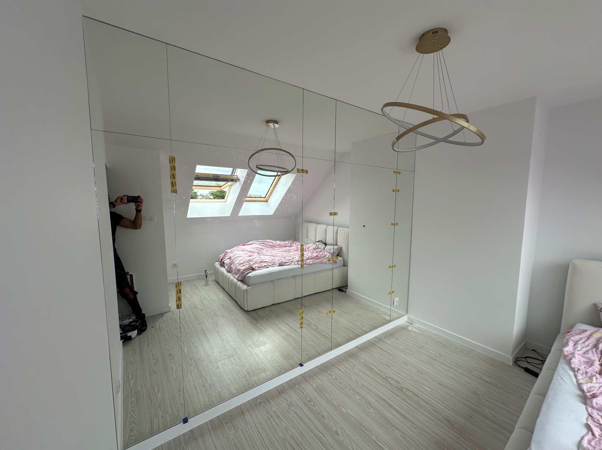 Duża, lustrzana szafa wnękowa z złotymi mocowaniami, odbijająca wnętrze sypialni z łóżkiem i oknem dachowym; widoczne odbicie fotografa.