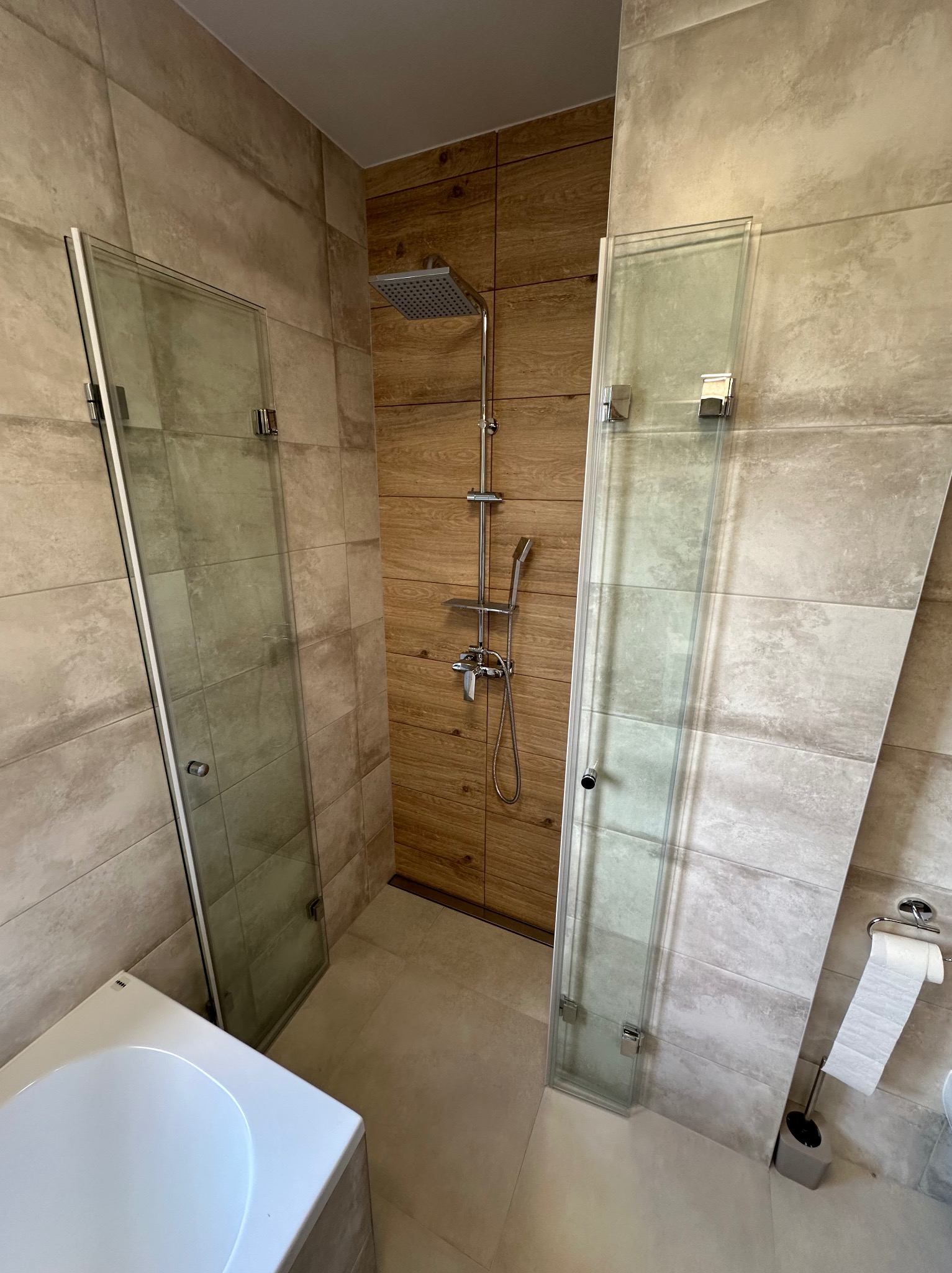 Nowoczesna kabina prysznicowa ze szklanymi drzwiami, wykończona płytkami w odcieniach beżu i drewna, z widoczną baterią prysznicową i toaletą w tle.