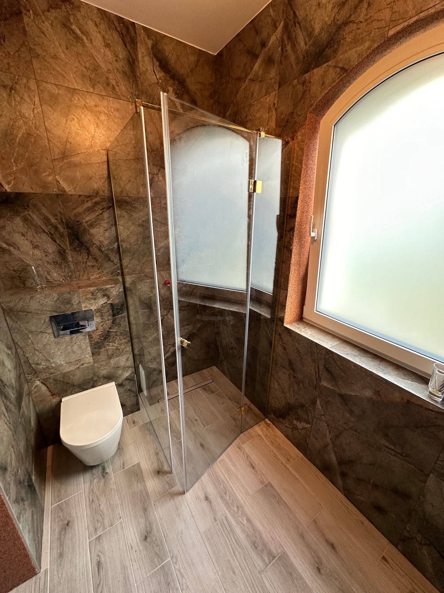 Wykończona łazienka z kabiną prysznicową ze szkła hartowanego, marmurowe płytki na ścianach, drewnopodobne płytki na podłodze, okno z zaokrąglonym górnym rogiem.