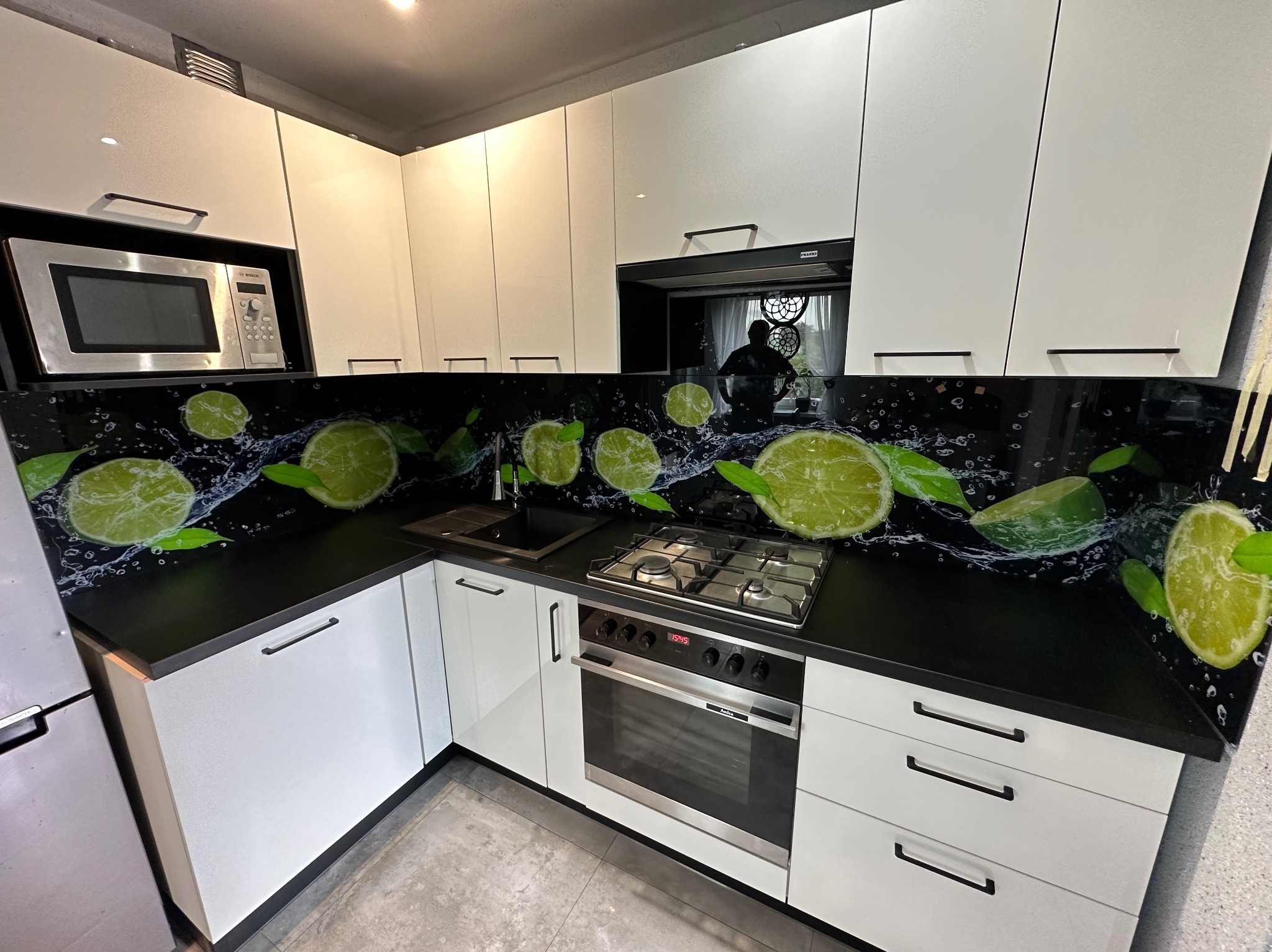 Nowoczesna kuchnia z białymi szafkami i czarnym blatem, na którym znajduje się panel szklany z motywem limonek i wody, odbicie osoby w okapie.
