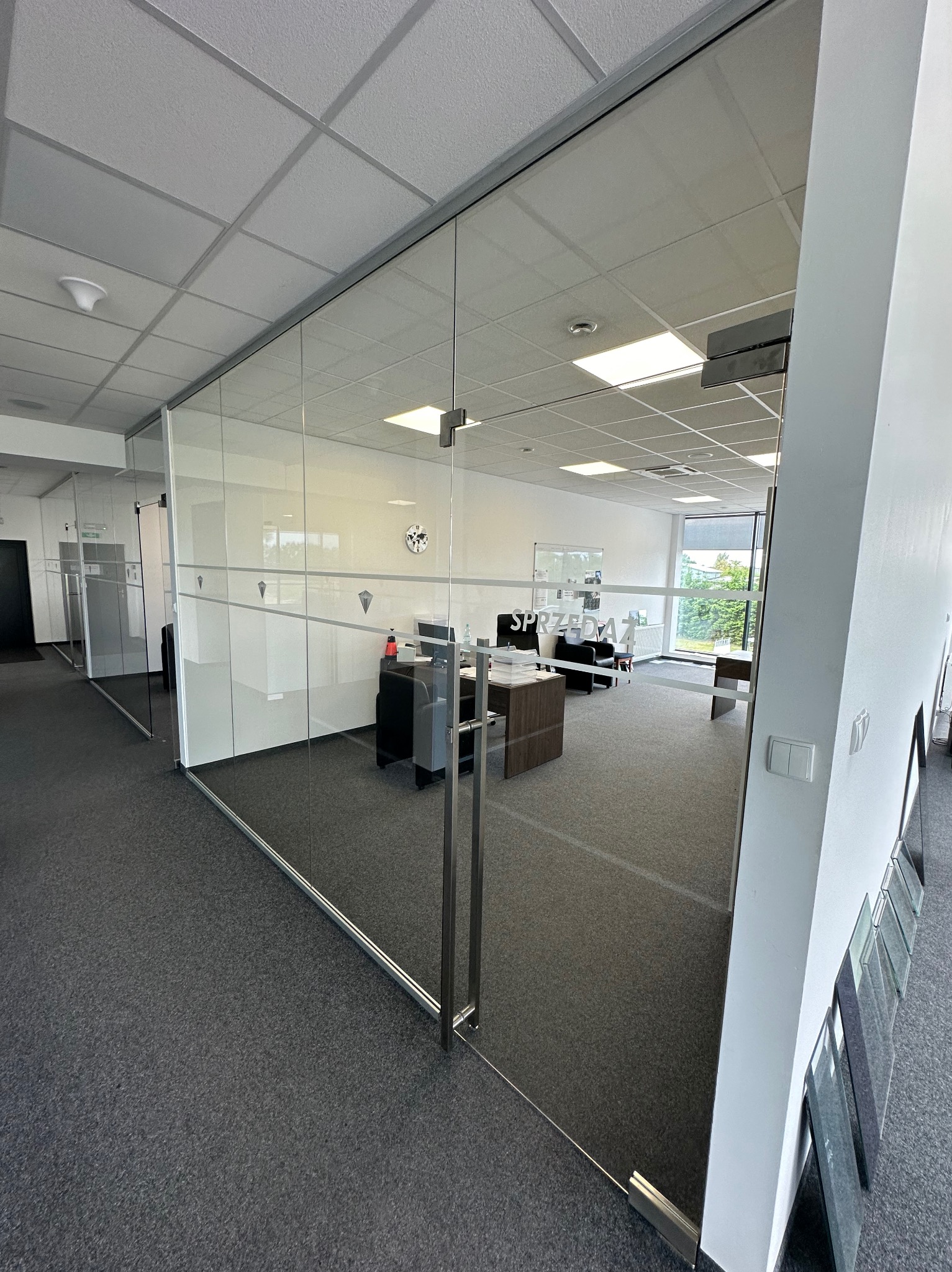 Szklana ściana działowa z drzwiami w biurze, widoczne biurka i krzesła w tle, na szkle delikatne wzory i napis 'Sprzedaż'.