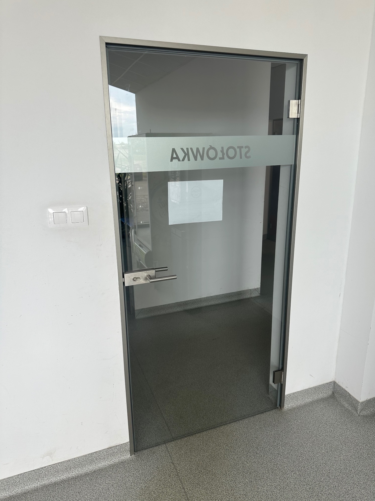 Szklane drzwi z metalową ramą i napisem na górnej części szyby, zamontowane w białej ścianie w pomieszczeniu biurowym.