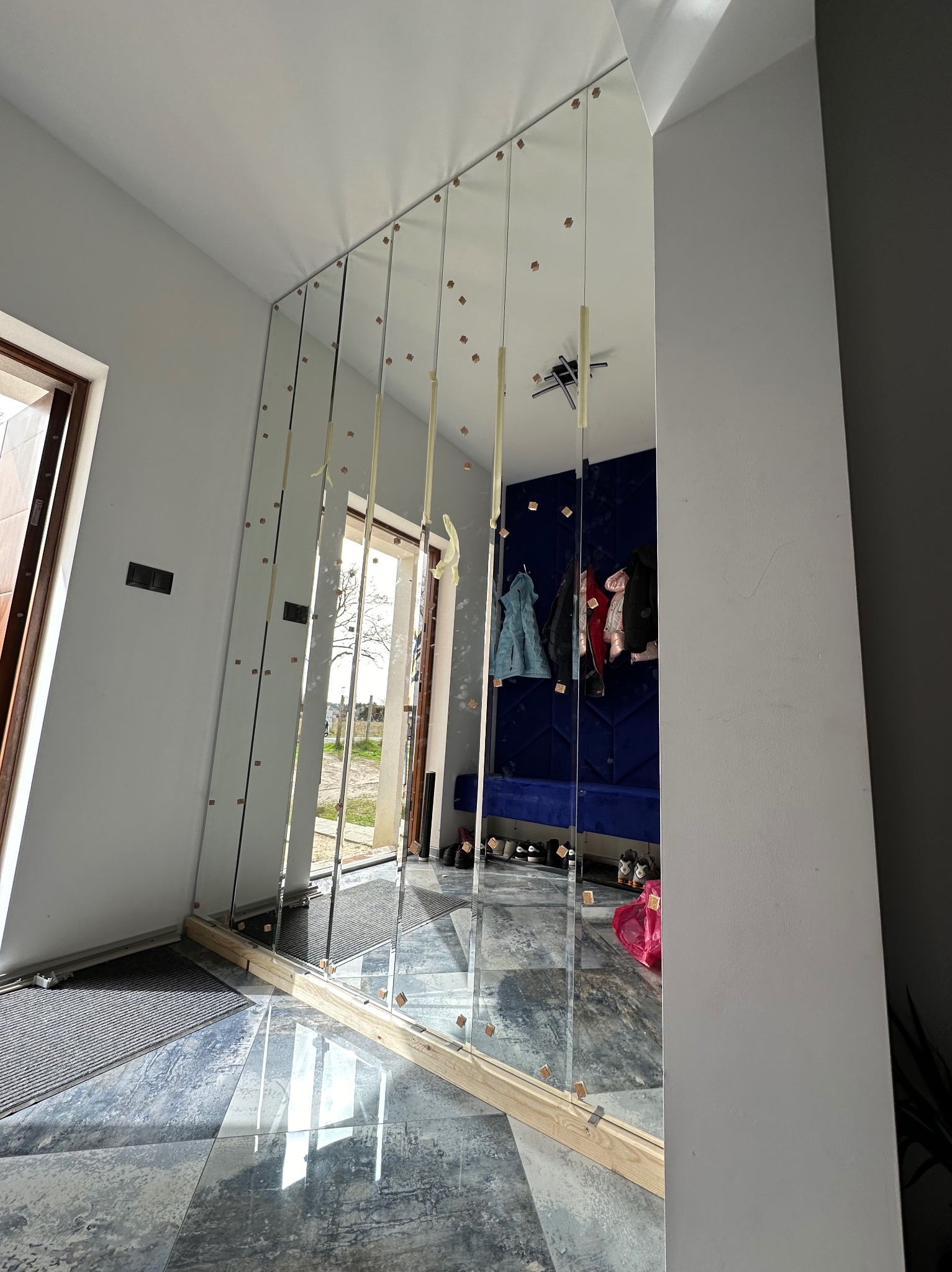 Montaż lustrzanej ściany z pionowych paneli, zabezpieczonych taśmą, w jasnym przedpokoju z widocznym wejściem i elementami garderoby w odbiciu.