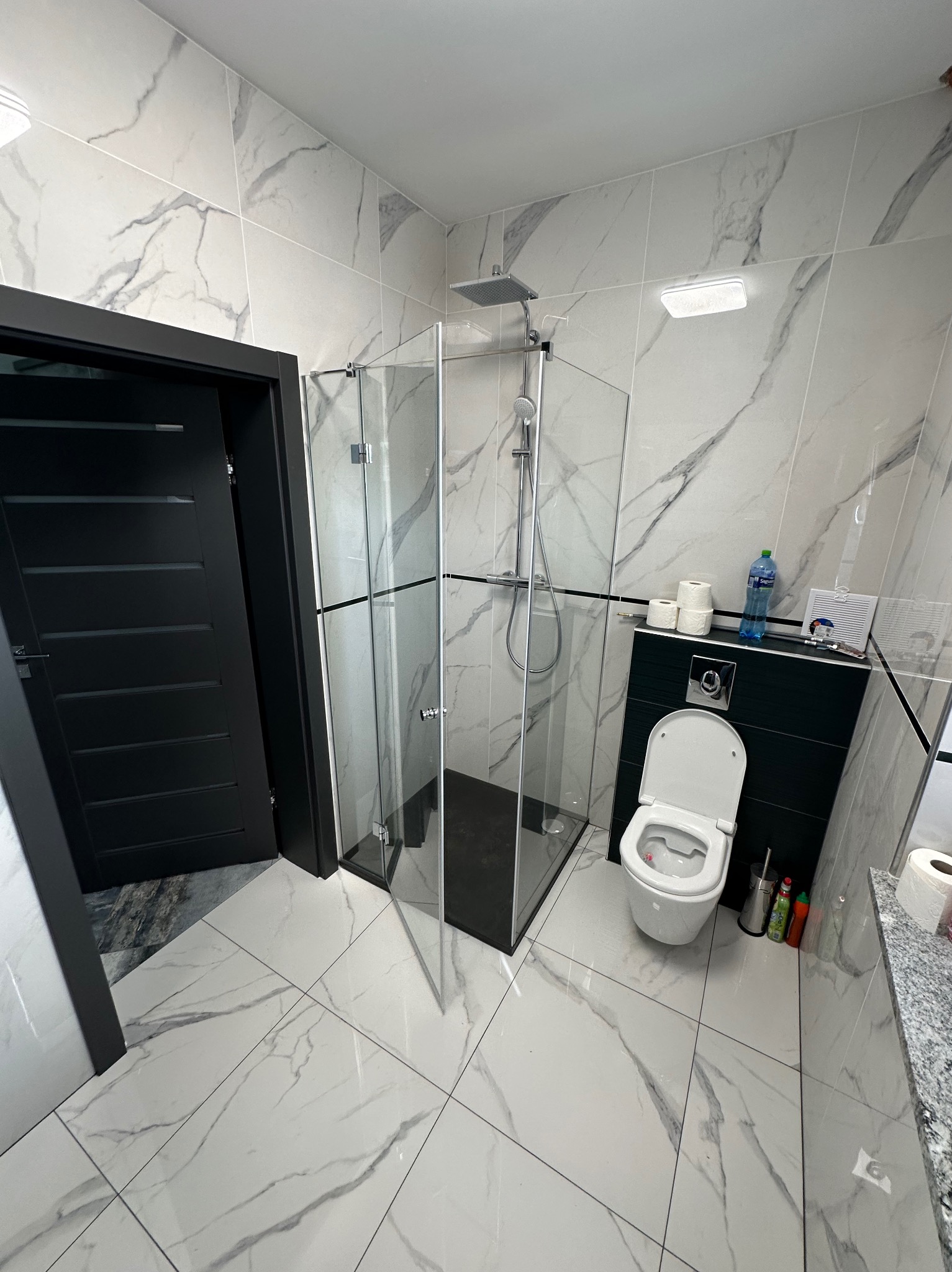 Nowoczesna łazienka z kabiną prysznicową ze szklanymi ścianami, czarną ramą i drzwiami otwieranymi na zewnątrz, białe marmurowe płytki na ścianach i podłodze, czarne drzwi wejściowe.