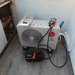 Klima Zbigniew Stolarek - Biała jednostka zewnętrzna klimatyzacji LG Dual Inverter, podłączona do pompy próżniowej Testo, z manometrami i wężami, ustawiona na szarych panelach podłogowych w rogu pomieszczenia.