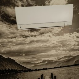 Klima Zbigniew Stolarek - Biała jednostka wewnętrzna klimatyzacji marki Mitsubishi zamontowana na ścianie z fototapetą przedstawiającą jezioro i góry w odcieniach szarości.