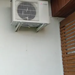 Biała jednostka zewnętrzna klimatyzacji Mitsubishi Electric zamontowana na białej ścianie budynku, widoczne drewniane elementy wykończeniowe dachu i ściany.