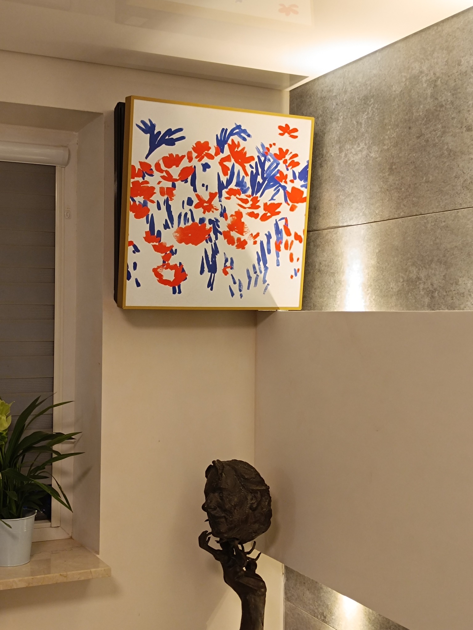 Wnętrze domu z obrazem w złotej ramie przedstawiającym abstrakcyjne kwiaty w kolorach czerwonym i niebieskim, rzeźba głowy na postumencie i roślina doniczkowa przy oknie.