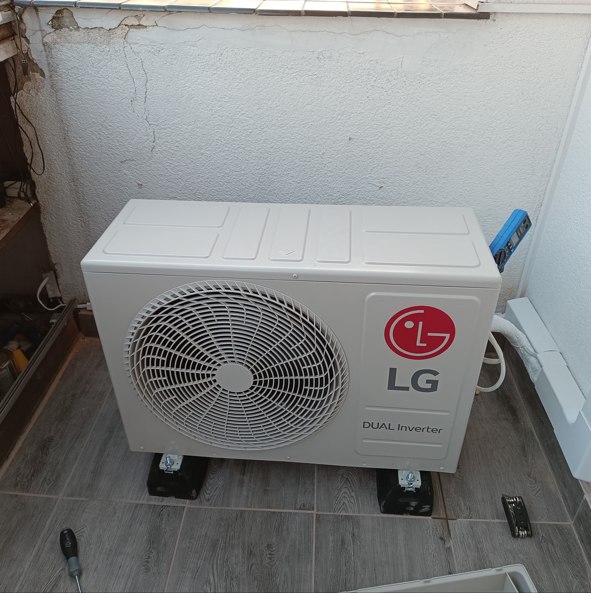Zewnętrzna jednostka klimatyzacji LG Dual Inverter na czarnych podkładkach, widoczne narzędzia instalacyjne, ściana z uszkodzeniami i miernik cyfrowy.