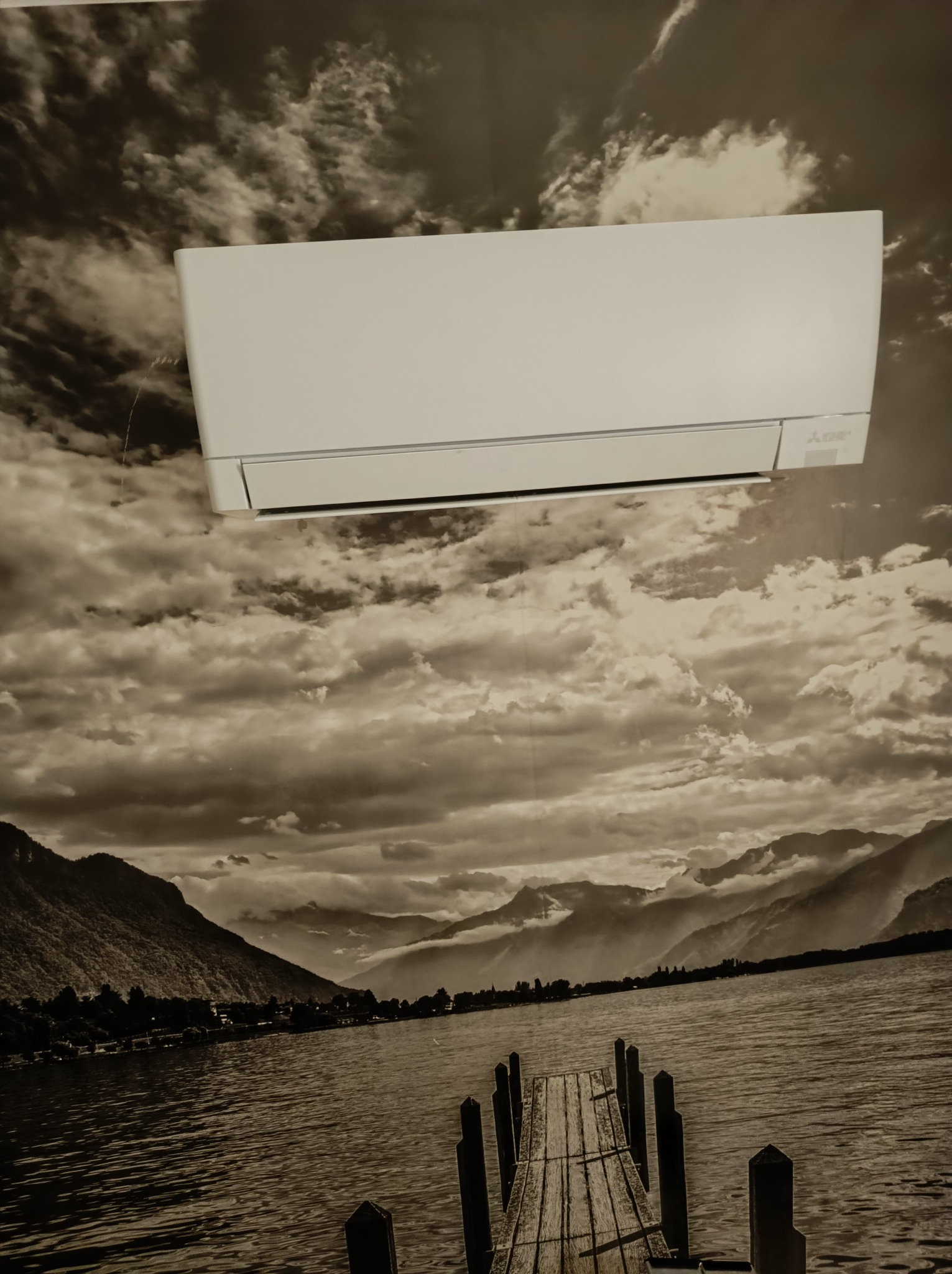 Biała jednostka wewnętrzna klimatyzacji marki Mitsubishi zamontowana na ścianie z fototapetą przedstawiającą jezioro i góry w odcieniach szarości.