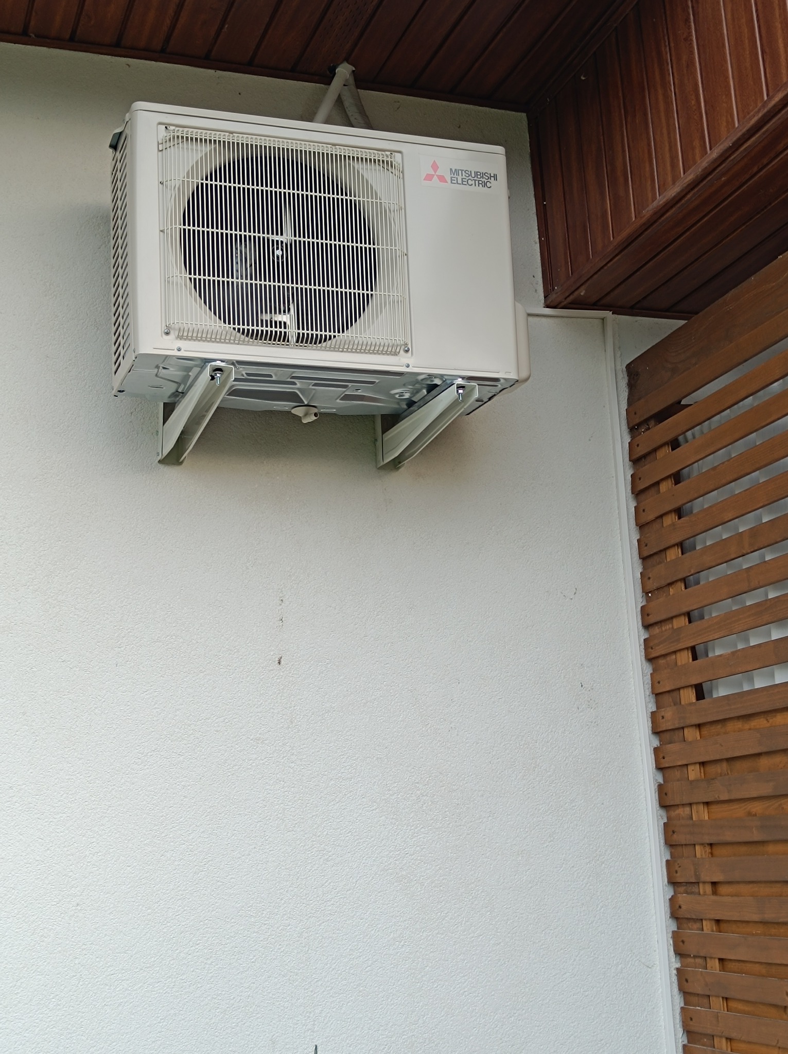 Biała jednostka zewnętrzna klimatyzacji Mitsubishi Electric zamontowana na białej ścianie budynku, widoczne drewniane elementy wykończeniowe dachu i ściany.