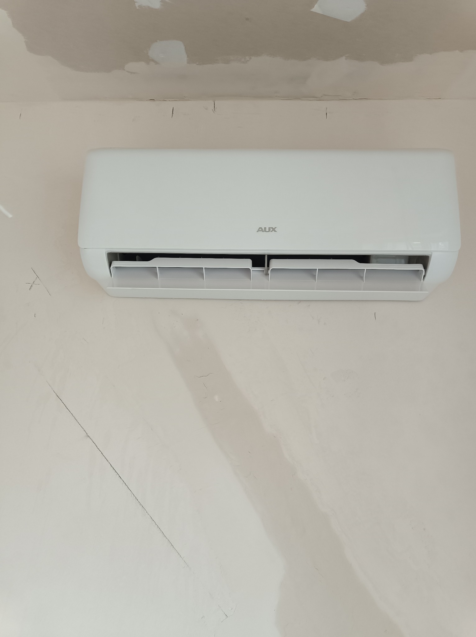 Biały klimatyzator marki AUX zamontowany na niedokończonej ścianie z widocznymi śladami szpachlowania i rysami.