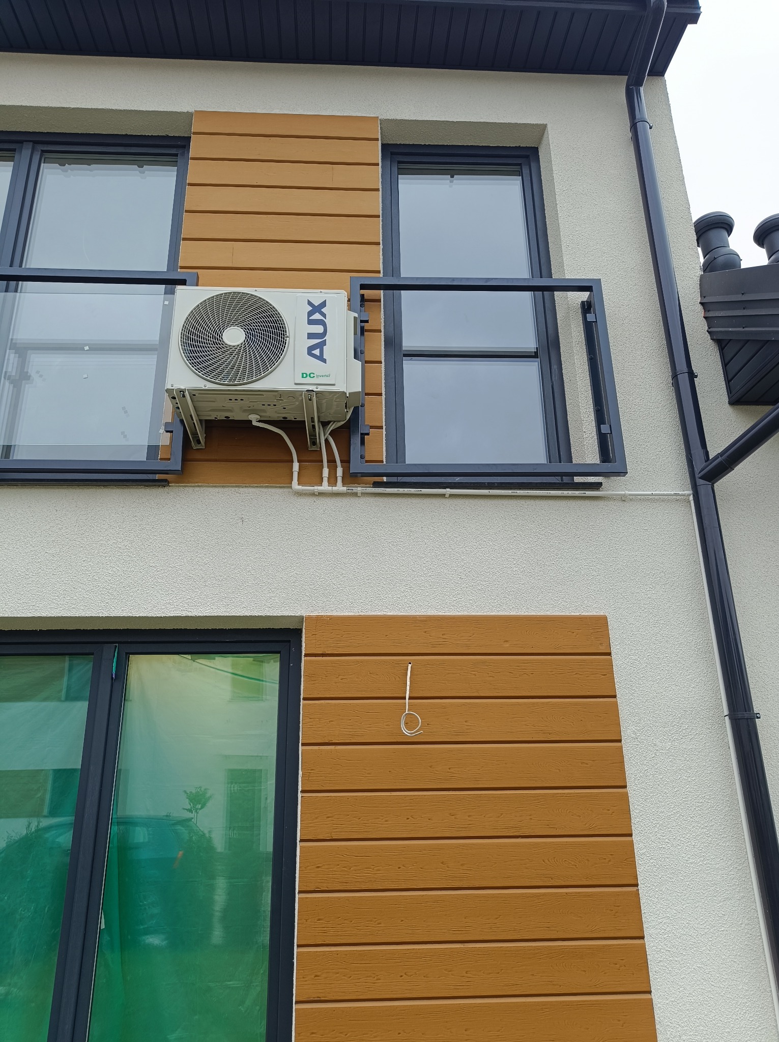 Biała jednostka klimatyzacyjna AUX zamontowana na elewacji domu z drewnianą okładziną, widoczne rury i kable, okna z czarnymi ramami i balkon z metalową balustradą.