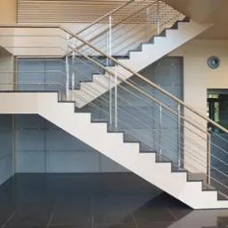 Nowoczesne schody dwubiegowe z drewnianymi stopniami i metalową balustradą w jasnym, minimalistycznym wnętrzu biurowym z panelami ściennymi i ciemną, błyszczącą podłogą.