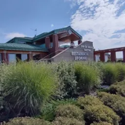 Budynek usługowy z zielonym dachem, ceglanymi elementami i napisem 'Restauracja Upominki', otoczony zielenią w słoneczny dzień.