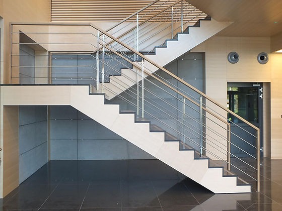 Nowoczesne schody dwubiegowe z drewnianymi stopniami i metalową balustradą w jasnym, minimalistycznym wnętrzu biurowym z panelami ściennymi i ciemną, błyszczącą podłogą.