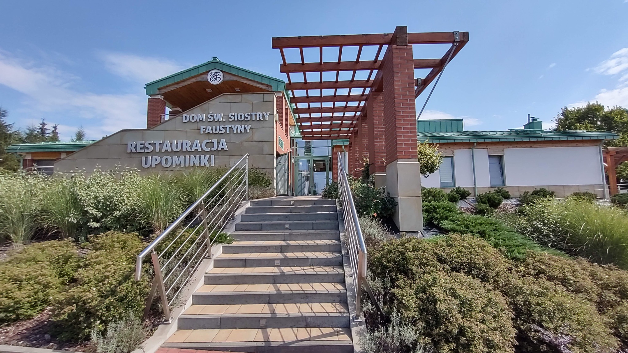 Fasada budynku z napisem 'Dom św. Siostry Faustyny Restauracja Upominki', schody wejściowe z metalową barierką, pergola nad wejściem, zieleń wokół