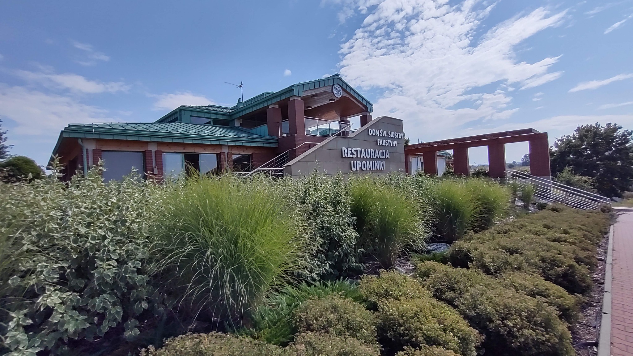 Budynek usługowy z zielonym dachem, ceglanymi elementami i napisem 'Restauracja Upominki', otoczony zielenią w słoneczny dzień.