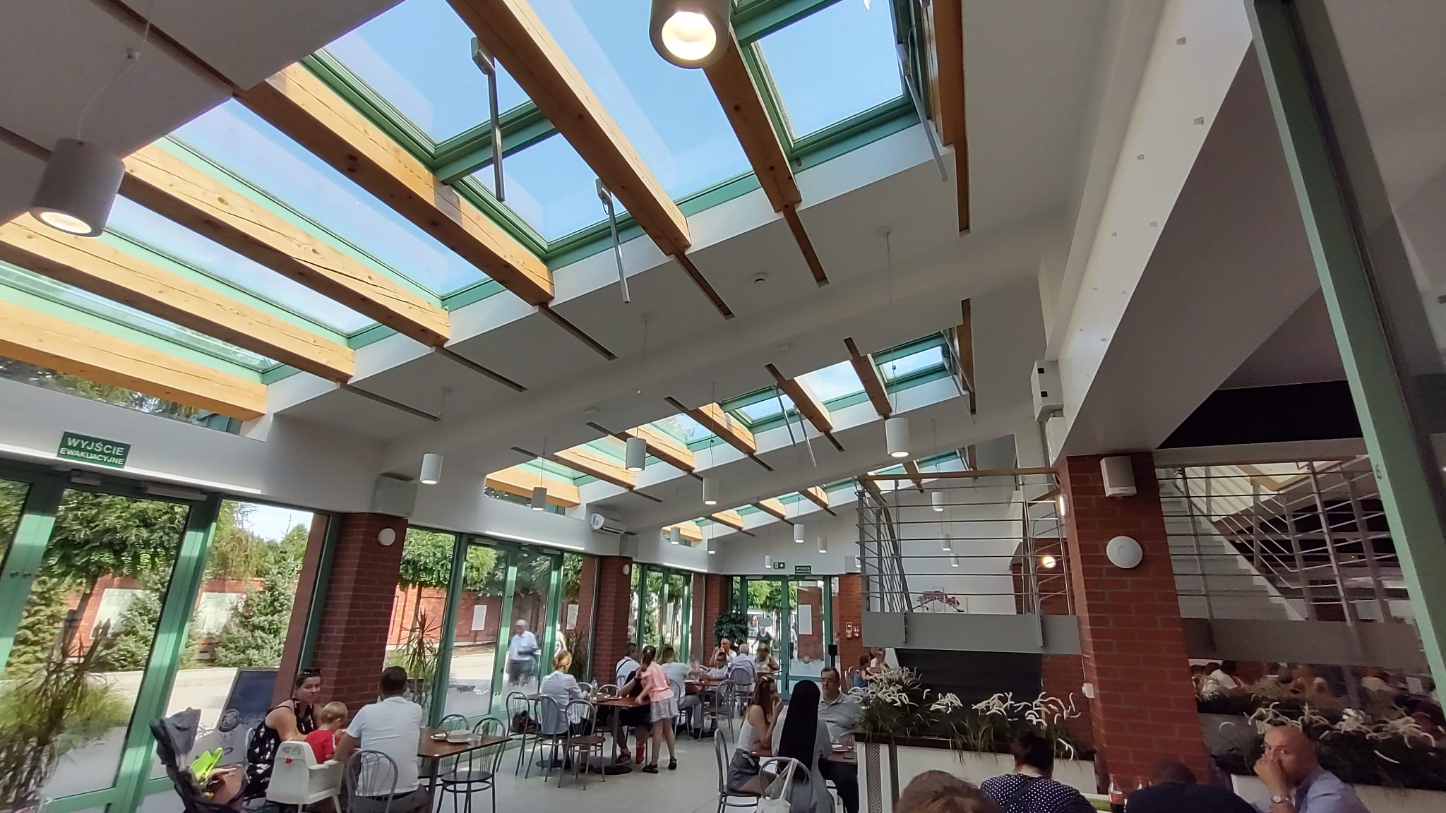 Wnętrze jasnej restauracji z przeszklonym dachem i drewnianymi belkami, widoczne stoliki z gośćmi i schody w tle.