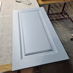 #Meble_Alex - Pomalowane na jasnoniebiesko drzwiczki szafki z frezowanymi ramkami leżą na warsztatowym stole, obok widać dokumenty, szmatkę i inne elementy wyposażenia warsztatu stolarskiego.