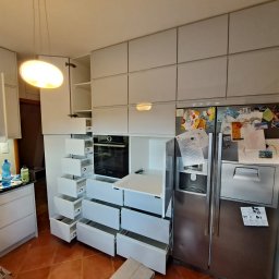 #Meble_Alex - Montaż białej kuchni na wymiar z otwartymi szufladami, widoczny piekarnik i lodówka z magnesami, osoba na drabince montuje górne szafki