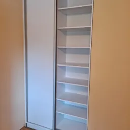 Biała szafa wnękowa z przesuwnymi drzwiami, widoczne puste półki, zamontowana w pomieszczeniu z beżowymi ścianami.