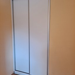 #Meble_Alex - Biała szafa wnękowa z przesuwnymi drzwiami i srebrnymi obramowaniami, ustawiona na panelach podłogowych przy beżowej ścianie.