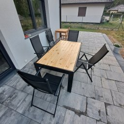 #Meble_Alex - Zewnętrzny stół z blatem z jasnego drewna i czarnymi metalowymi nogami, otoczony składanymi krzesłami na szarym brukowanym tarasie przy domu z szarą elewacją.