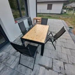 Zewnętrzny stół z blatem z jasnego drewna i czarnymi metalowymi nogami, otoczony składanymi krzesłami na szarym brukowanym tarasie przy domu z szarą elewacją.