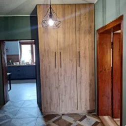 Trzydrzwiowa szafa z drewnopodobnym fornirem i czarnymi, pionowymi uchwytami, oświetlona wiszącą lampą z geometrycznym kloszem, w tle widoczny fragment kuchni.